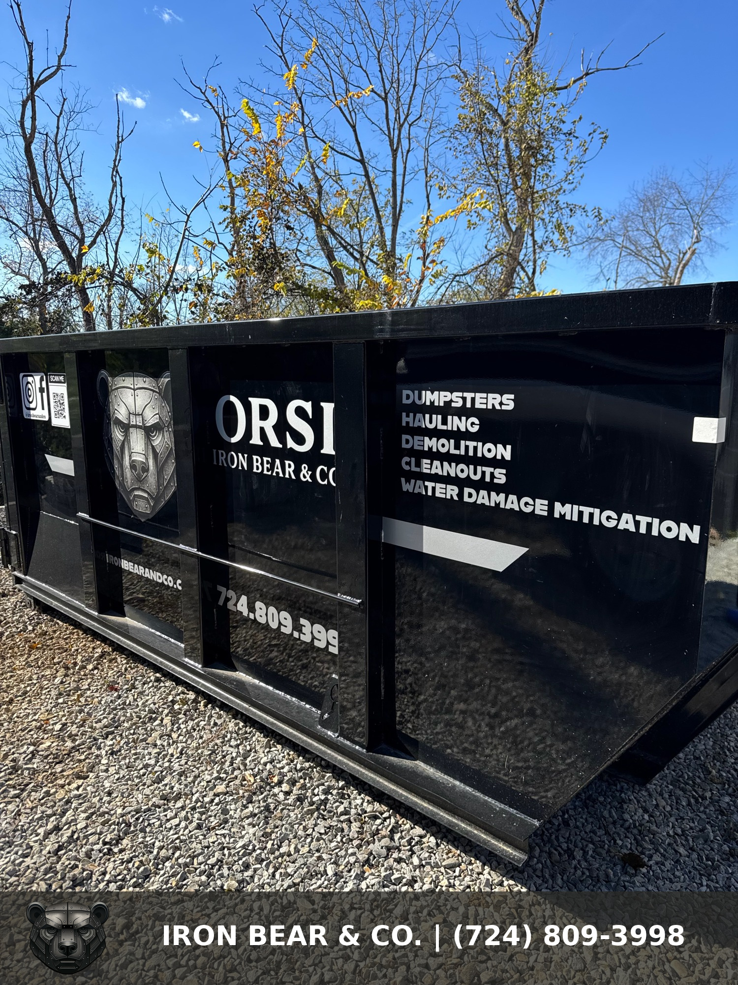 Black Iron Bear & Co. roll-off dumpster lettered for dumpsters, hauling, demolition, and cleanouts, parked on gravel with fall trees behind