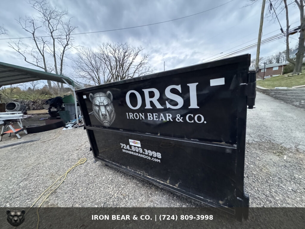 Black Iron Bear & Co. roll-off dumpster with the bear logo and phone number, set on a gravel drive under bare late-fall trees