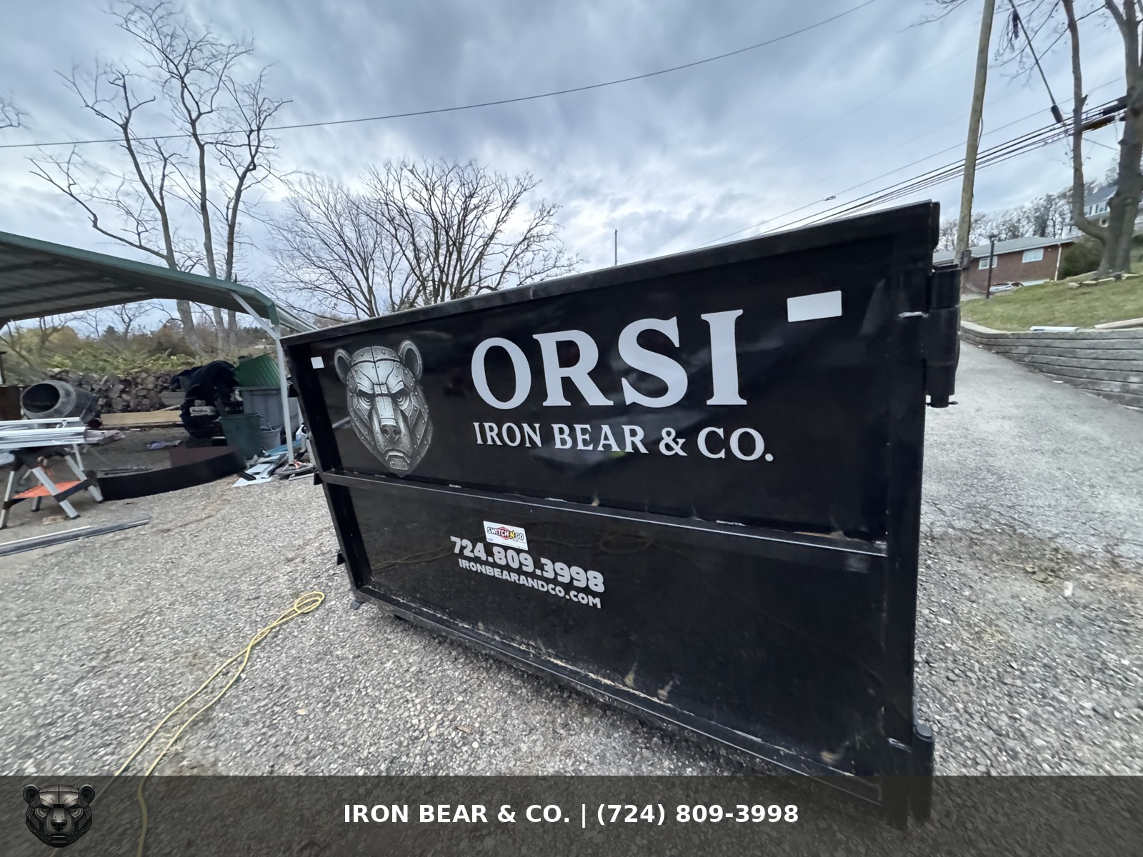 Black Iron Bear & Co. roll-off dumpster with the bear logo and phone number, set on a gravel drive under bare late-fall trees
